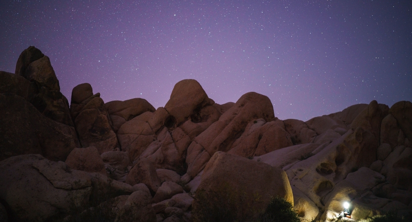 A headlamp illuminates a person sitting at the bottom of a large rock formation, resting under a purple night sky dotted with stars